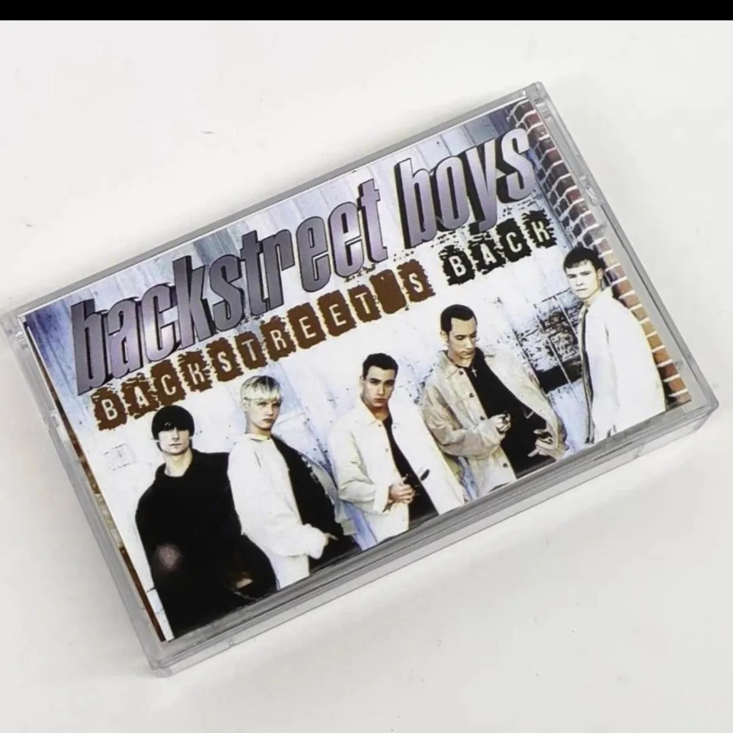 Audio Music Of - เทปคาสเซ็ตเสียงเพลงป๊อปอะนาล็อกวินเทจ กลุ่มบอยแบนด์ฮาร์โมนี แทร็กเพลงย้อนยุค ปกภาพบุคคลบนกำแพงอิฐ ดนตรีกายภาพที่ชวนให้คิดถึงอดีต